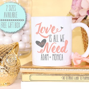 May include: White ceramic coffee mug with a pink and gray design. The mug says "Love is all we need" with a heart and the date "05.18.15". Below the text is "Adam + Monica".