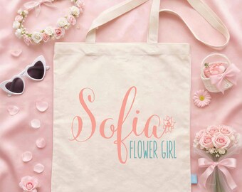 Puede incluir: Una bolsa de tela color crema con el nombre Sofia en letra cursiva coral grande y las palabras FLOWER GIRL en letras mayúsculas de color turquesa. La bolsa está rodeada por una corona de flores, gafas de sol en forma de corazón y un ramo.
