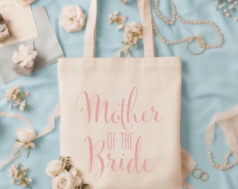 May include: A cream-coloured canvas tote bag featuring the text Mother of the Bride printed in a pink cursive script. The bag has two matching fabric handles and a rectangular shape, presented as a flat lay item with a clean, simple design.