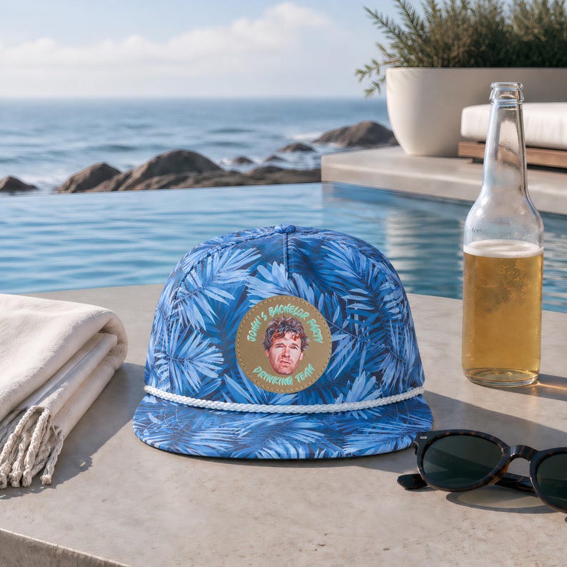 Op de afbeelding: Een blauwe honkbalpet met een palmbladpatroon, een wit koord en een ronde beige patch aan de voorzijde. De patch bevat een portret van een persoon en de tekst "JONH'S BACHELOR PARTY DRINKING TEAM" in een cirkelvormige opstelling.
