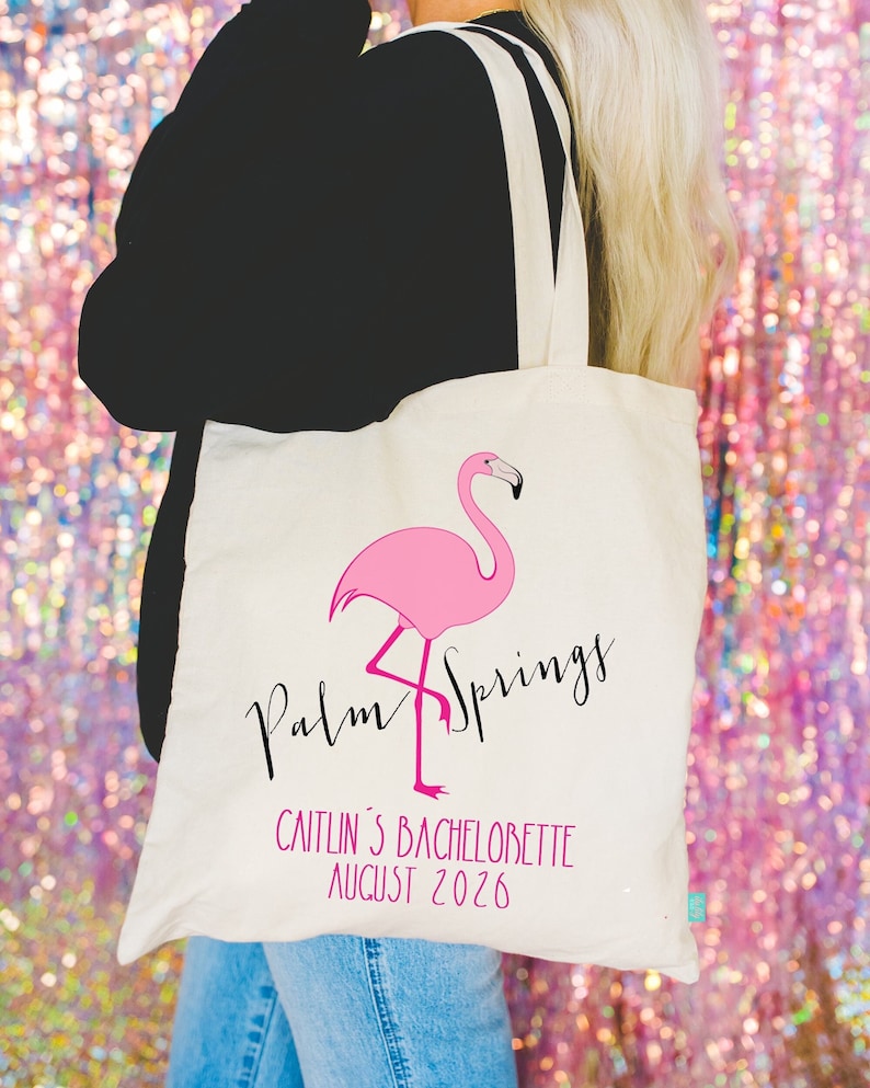 May include: A white canvas tote bag with a pink flamingo illustration and the text "Palm Springs" in black script. The bag also has the text "Caitlin's Bachelorette August 2026" printed on it.