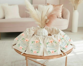 May include: A collection of off-white drawstring fabric bags arranged in a circle on a round table. Each bag features a printed floral wreath design in pink and green, with the text "Jackie + Steven" and "06.07.202X" and "HANGOVER KIT".
