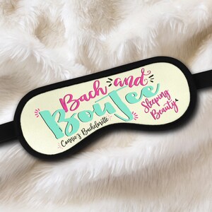 May include: A white sleep mask with black elastic straps. The mask has the text "Bach and Boujee" in pink and teal lettering. The text "Connie's Bachelorette" is written in black below the main text. The text "Sleeping Beauty" is written in pink on the right side of the mask.