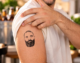 Op de afbeelding: Een tijdelijke tatoeage met een zwarte inkttekening van een persoon met kort haar en een volle, dikke baard. Het ontwerp is aangebracht op de bovenarm en toont gedetailleerde gelaatstrekken en donkere arceringen in de baard.
