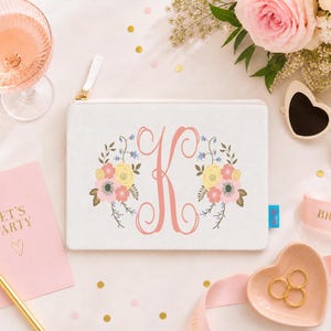 May include: A white rectangular zipped pouch featuring a large coral-coloured script letter K in the centre, flanked by floral illustrations in shades of yellow, pink, and blue. The pouch has a white zip pull and a small blue side tag.