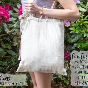 May include: A white canvas tote bag with pink ribbon handles. The bag is 15 inches wide and 16 inches high. The text on the bag reads: "Tote Bags: Measure 15"W x 16"H". The text on the right side of the bag reads: "Can Easily Hold: 4 Bottles of Wine, A Rolled Up Beach Towel, Sun Tanning Lotion, A Bottle of Water, A Magazine + More!"