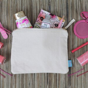 May include: A white canvas zippered pouch with a gold zipper, filled with various pink and white beauty products, including a bottle of hand cream, a small bottle of lotion, a lipstick, a nail polish, a blush compact, and a pencil. The pouch is sitting on a wooden surface.
