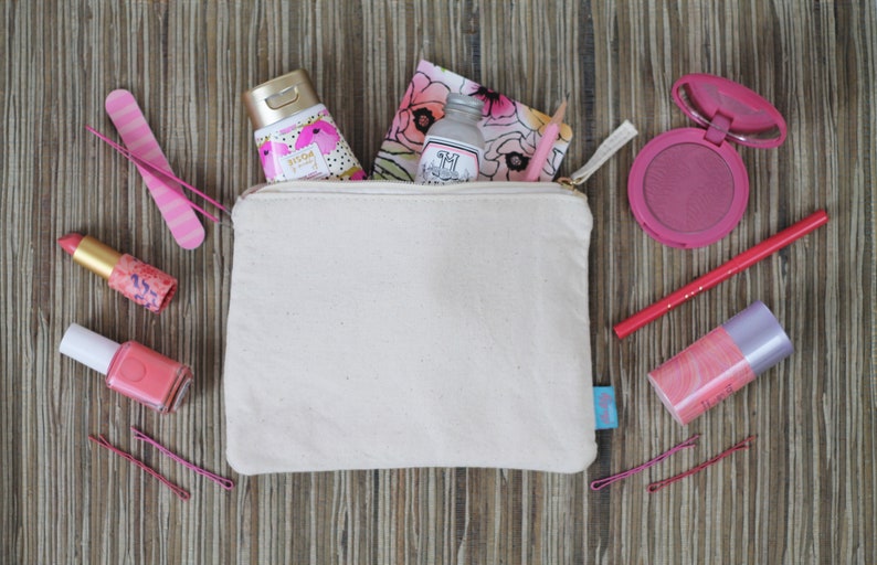 May include: A white canvas zippered pouch with a gold zipper, filled with various pink and white beauty products, including a bottle of hand cream, a small bottle of lotion, a lipstick, a nail polish, a blush compact, and a pencil. The pouch is sitting on a wooden surface.