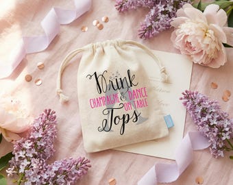 May include: A small off-white drawstring pouch featuring the black and pink text Drink CHAMPAGNE & DANCE ON TABLE Tops, with a graphic of two champagne flutes. The pouch is surrounded by light purple lilac flowers and a light purple ribbon.