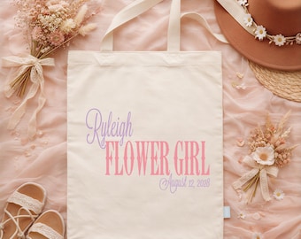 Puede incluir: Una bolsa de tela color crema con el texto morado Ryleigh, el texto rosa FLOWER GIRL y el texto morado August 12, 2028. La bolsa tiene dos asas y se muestra junto a un sombrero, flores secas y un par de sandalias.