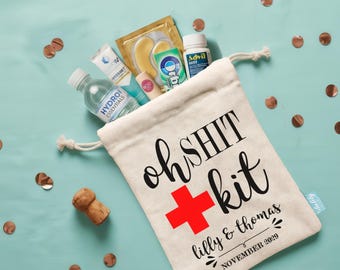 May include: A cream-coloured drawstring canvas bag printed with the text "oh SHIT kit", a red cross symbol, and "lilly & thomas NOVEMBER 2029". The bag is filled with a water bottle, a tube of lotion, gold eye patches, gum, and a bottle of Advil.