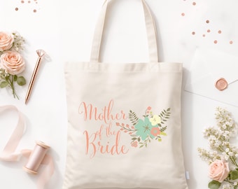 May include: A cream-coloured canvas tote bag featuring the text Mother of the Bride in a coral script font, accompanied by a floral illustration in shades of teal, coral, and yellow, with two sturdy shoulder straps and a rectangular body shape.