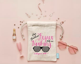 May include: A beige drawstring canvas pouch featuring the text "Aug's Bachelorette Regret Nothing July 2027 Hangover Kit" in black and pink, with a graphic of black and white striped sunglasses. Includes a mini champagne bottle and heart-shaped glasses.