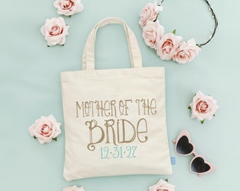 May include: A cream-coloured canvas tote bag featuring the brown text MOTHER OF THE BRIDE and the teal date 12.31.27, surrounded by five individual dusty pink artificial roses, a dusty pink floral crown, and heart-shaped sunglasses.