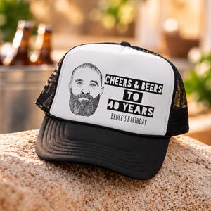 May include: A black and white trucker hat featuring a graphic illustration of a bearded man and the black text Cheers & Beers to 40 Years Bruce's Birthday printed on the white front panel, with a black mesh back and a solid black brim.