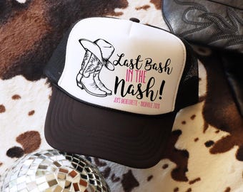 Könnte beinhalten: Eine schwarz-weiße Trucker-Cap mit braunem Schirm. Das Vorderteil zeigt eine Grafik eines Cowboystiefels und eines Hutes mit dem Text "Last Bash IN THE Nash!" in Schwarz und Pink sowie "JEN'S BACHELORETTE • NASHVILLE 2028" in kleiner pinker Schrift.