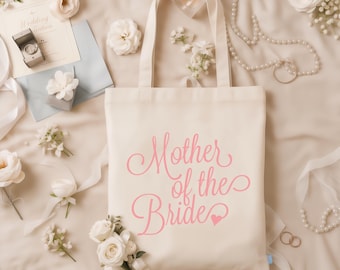 May include: A cream-coloured canvas tote bag featuring the pink script text Mother of the Bride with a small heart icon. The bag has two shoulder straps and is displayed with white flowers, pearl necklaces, and a wedding invitation card.