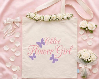 Puede incluir: Una bolsa de tela color crema con el texto Chloé Flower Girl en letra cursiva rosa y tres mariposas moradas. La bolsa está adornada con una hilera de rosas artificiales blancas en el borde superior e incluye un lazo de cinta blanca a juego.