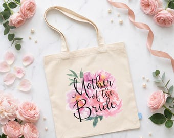 May include: A cream-coloured canvas tote bag featuring a watercolour illustration of pink peonies and the black script text Mother of the Bride. The bag has two shoulder straps and a small blue fabric tag on the side seam, presented in a flat layout.