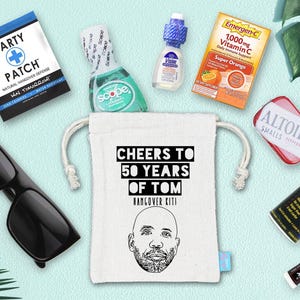 May include: An assortment of items, including sunglasses, a drawstring bag with the text "CHEERS TO 50 YEARS OF TOM", and various small health and wellness products. The items are arranged on a light blue surface.