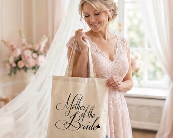 May include: A cream-coloured canvas tote bag featuring the black script text Mother of the Bride with a small heart icon. The bag has two fabric handles and a rectangular shape, constructed from a light-coloured woven textile material.