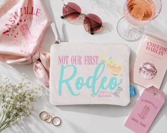May include: A beige canvas zipper pouch featuring the text "NOT OUR FIRST Rodeo" in pink and teal, with a yellow guitar graphic labeled "Nashville 05.18.28" and a small cowboy boot icon labeled "Sarah's Bachelorette" printed on the front.