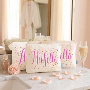 May include: Three off-white canvas zipped pouches featuring a pink script name Michelle and scattered gold and pink confetti dots. The pouches are arranged on a marble surface alongside a glass perfume bottle, a pink rose, and a champagne flute.