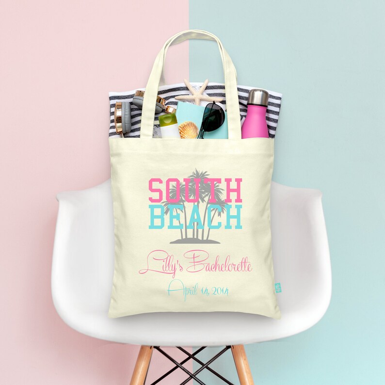 May include: A white canvas tote bag with the text "South Beach" in pink and blue lettering. The bag also has the text "Lilly's Bachelorette April 18, 2014" in gray lettering. The bag is filled with various items, including a beach towel, sunglasses, a water bottle, and a seashell.