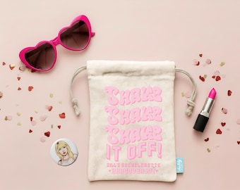 May include: A cream-colored drawstring pouch featuring the pink text "SHAKE SHAKE SHAKE IT OFF!" and "DILL'S BACHELORETTE HANGOVER KIT". The pouch is accompanied by heart-shaped pink sunglasses, a tube of pink lipstick, and a circular pin with a blonde illustration.