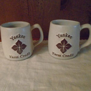 May include: Two off-white ceramic mugs with handles. Each mug features the words "Yankee Farm Credit" and a brown floral design. The mugs are on a white surface with a wooden background.