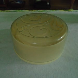 May include: A round, pale yellow plastic container with a matching lid. The lid features a raised, embossed swirl pattern across its surface. The container has smooth, vertical sides and a flat base, presenting a simple, functional design.