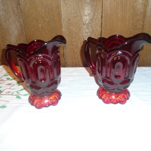 May include: Two red glass creamer pitchers with a textured design. The pitchers have a scalloped rim and a footed base.