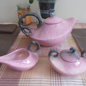 May include: A pink speckled ceramic tea set, including a teapot with a lid, a creamer, and a sugar bowl with a lid. The handles and accents are a dark gray-blue color. The set is displayed on a patterned tablecloth.
