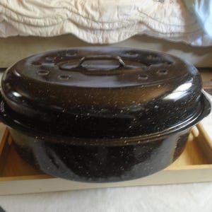 May include: A black speckled roasting pan with a lid and handles. The pan is sitting on a wooden tray. The lid has several small holes and a handle. This cookware is designed for roasting meats and vegetables.