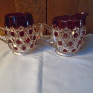May include: A vintage set of a red glass creamer and sugar bowl with a diamond pattern and red accents.