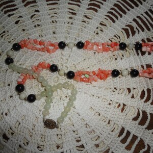 Puede incluir: Un collar de varias hebras con coral rosa, cuentas blancas y negras. El collar est&aacute; sobre una superficie de crochet blanco.