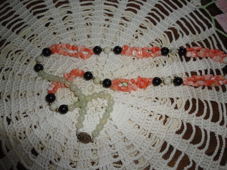 Puede incluir: Un collar de varias hebras con coral rosa, cuentas blancas y negras. El collar est&aacute; sobre una superficie de crochet blanco.