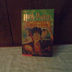 May include: A hardcover book titled "Harry Potter and the Goblet of Fire" by J.K. Rowling. The cover features a smiling boy with glasses, and two other figures. The book has a green and gold color scheme with the word "Scholastic" at the bottom.