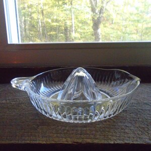 May include: A clear glass citrus juicer with a ridged bowl and a pointed center section. The juicer is designed to hold a citrus fruit and squeeze the juice into the bowl.