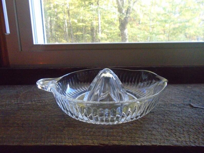 May include: A clear glass citrus juicer with a ridged bowl and a pointed center section. The juicer is designed to hold a citrus fruit and squeeze the juice into the bowl.