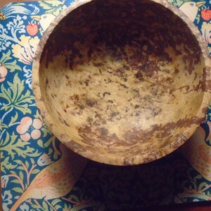 May include: A round, light-colored wooden bowl with dark brown and tan markings. The bowl sits on a blue patterned surface with floral and bird designs. The bowl's interior shows signs of wear.