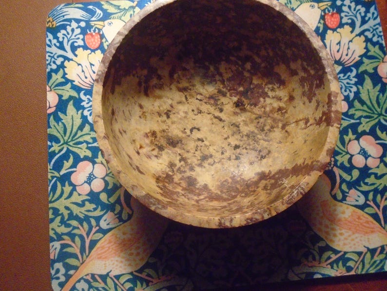 May include: A round, light-colored wooden bowl with dark brown and tan markings. The bowl sits on a blue patterned surface with floral and bird designs. The bowl's interior shows signs of wear.