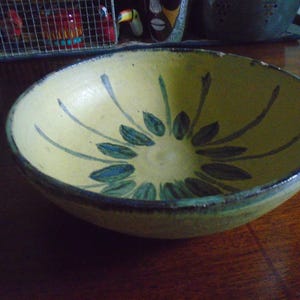 May include: A shallow, round ceramic bowl with a pale yellow glaze and a hand-painted floral motif in dark green and blue. The interior features a central cluster of petal-like shapes with radiating lines, and the rim has a dark, weathered edge.