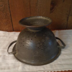 May include: A vintage metal colander with a dark, aged appearance. The colander features a bowl-shaped base with numerous small holes, a flared rim, and two curved handles. It has a smaller, raised bowl on top.