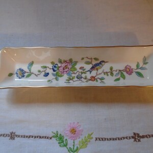 May include: A rectangular porcelain tray with a white base and a gold-trimmed scalloped edge. The center features a colorful botanical illustration of pink and blue flowers, green leaves, and a small blue and yellow bird perched on a branch.