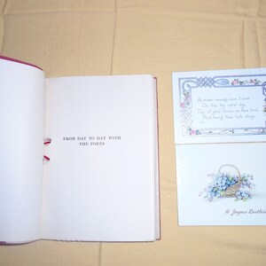 May include: An open book titled "From Day to Day with the Poets" with a red ribbon bookmark. Two greeting cards are visible, one with a floral design and the text "A tender message, dear, I send On this thy natal day They all good fairies on thee tend, And bring thee back alway", and the other with a basket of blue flowers and the text "A Joyous Birthday Greeting".