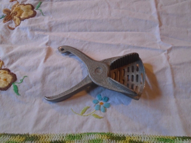 May include: A silver metal citrus juicer with a handle and a gridded base. The juicer is on a white tablecloth with embroidered flowers.