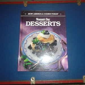 Może przedstawiać: Książka kucharska zatytułowana "Woman's Day Desserts" z fioletową okładką i zdjęciem deseru z bor&oacute;wkami i bitą śmietaną na białym talerzu.
