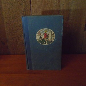 May include: A blue hardcover book with a circular illustration on the front cover. The illustration depicts a young girl in a red dress standing on a building with a man in a green suit behind her.
