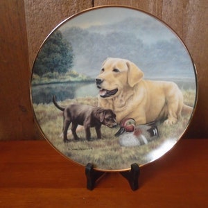 May include: A porcelain plate with a gold rim features a painting of a yellow Labrador Retriever and a chocolate Labrador Retriever puppy. The dogs are in a grassy field with a duck in the foreground. The plate is on a black stand.